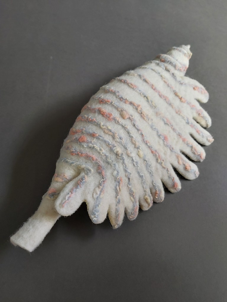 A white wet felted conch shell with ribs in pale grey and peach.